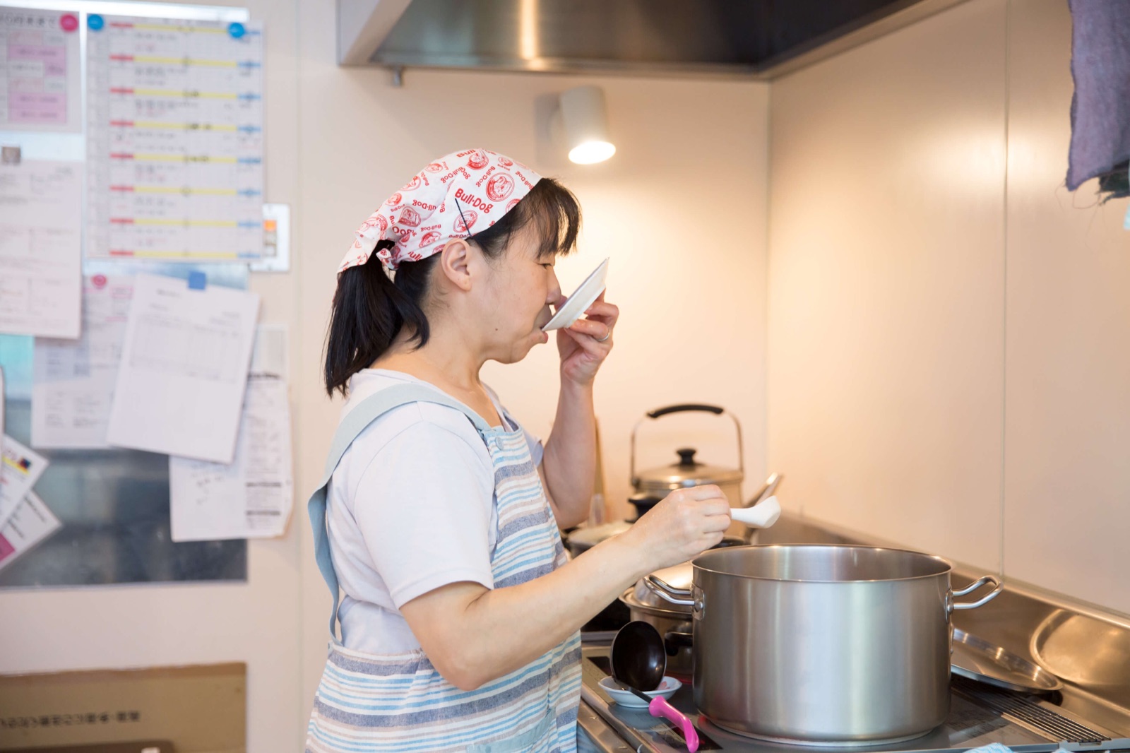 コンロの前で小皿から味見をするエプロン姿の女性