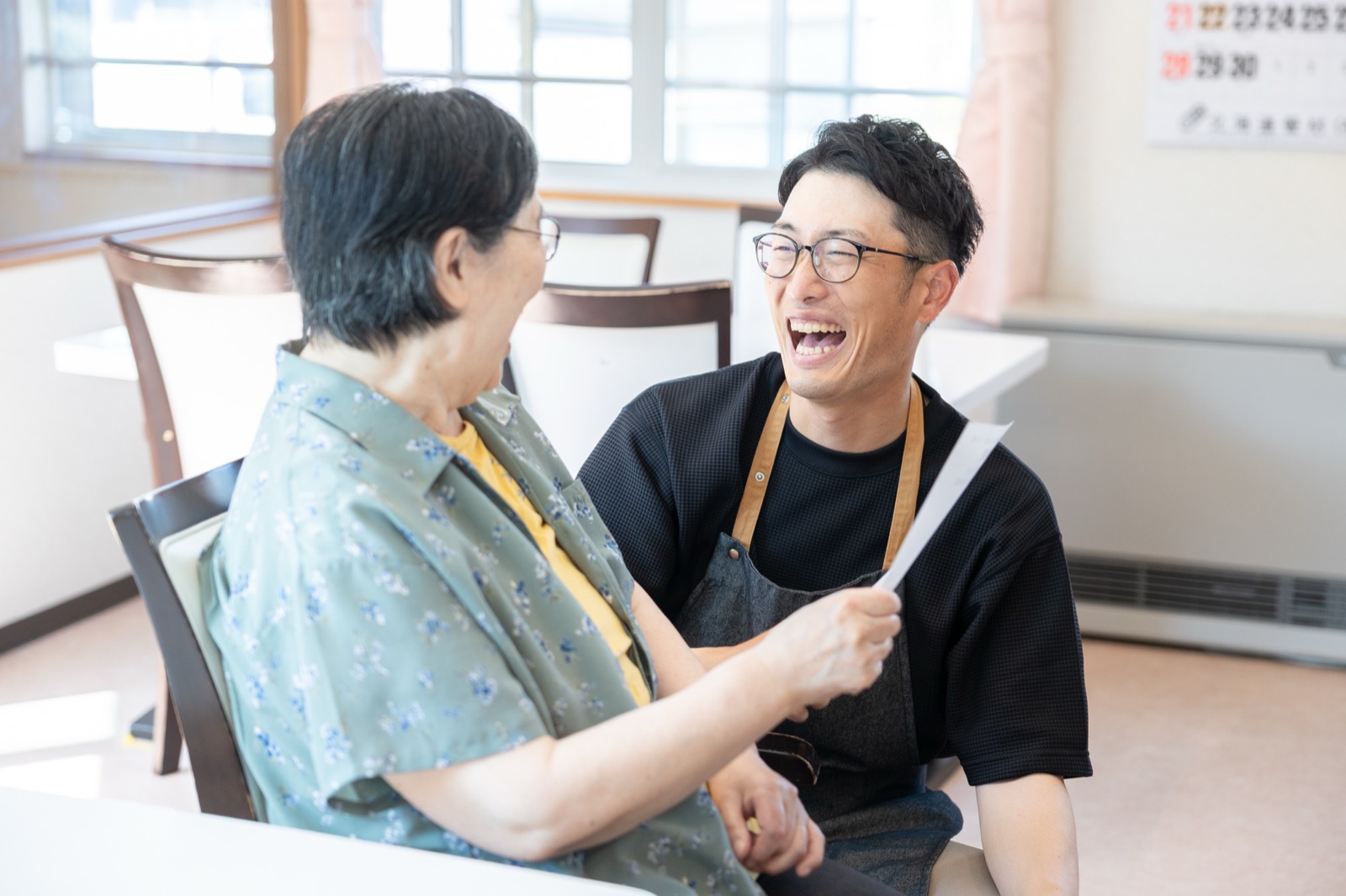 椅子に座り、紙を手に笑い合う高齢の女性とエプロン姿の男性